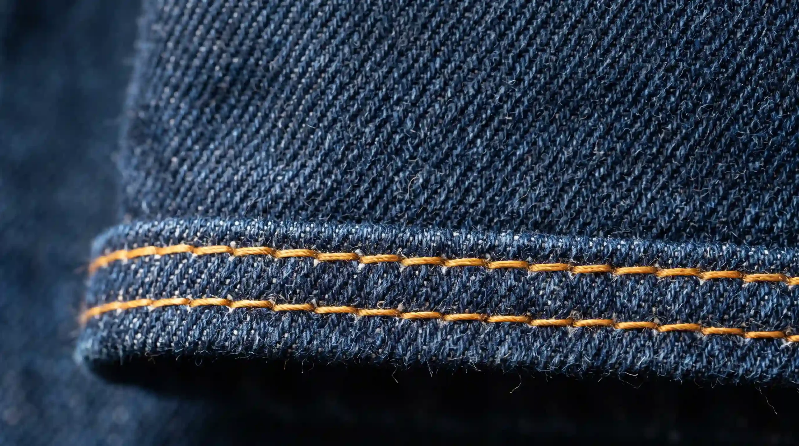 Macro photography focused on the diagonal twill texture and double-stitch reinforcement of heavy 14-ounce dark indigo denim.