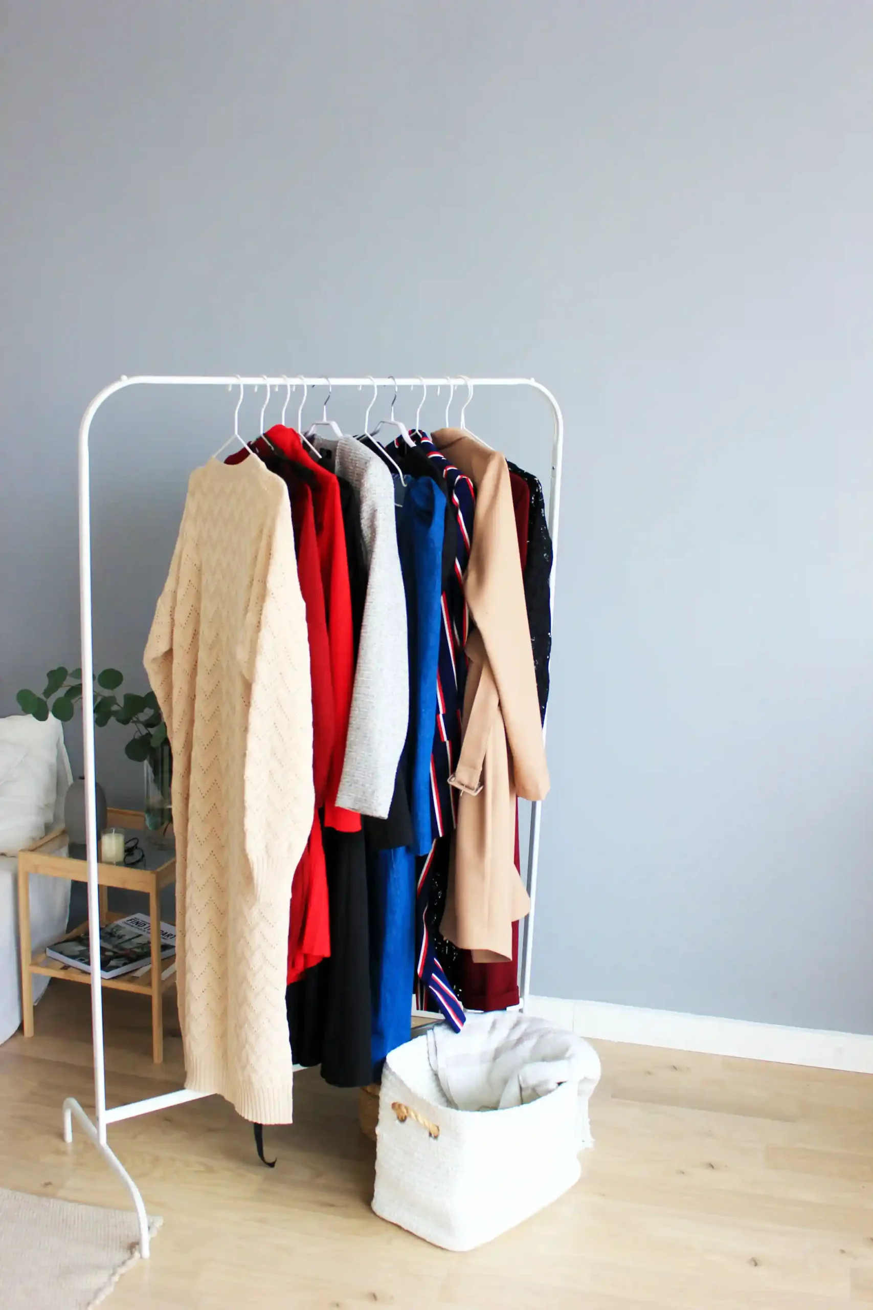 A white clothing rack holding cream, red, grey, blue, and camel garments against a pale grey wall