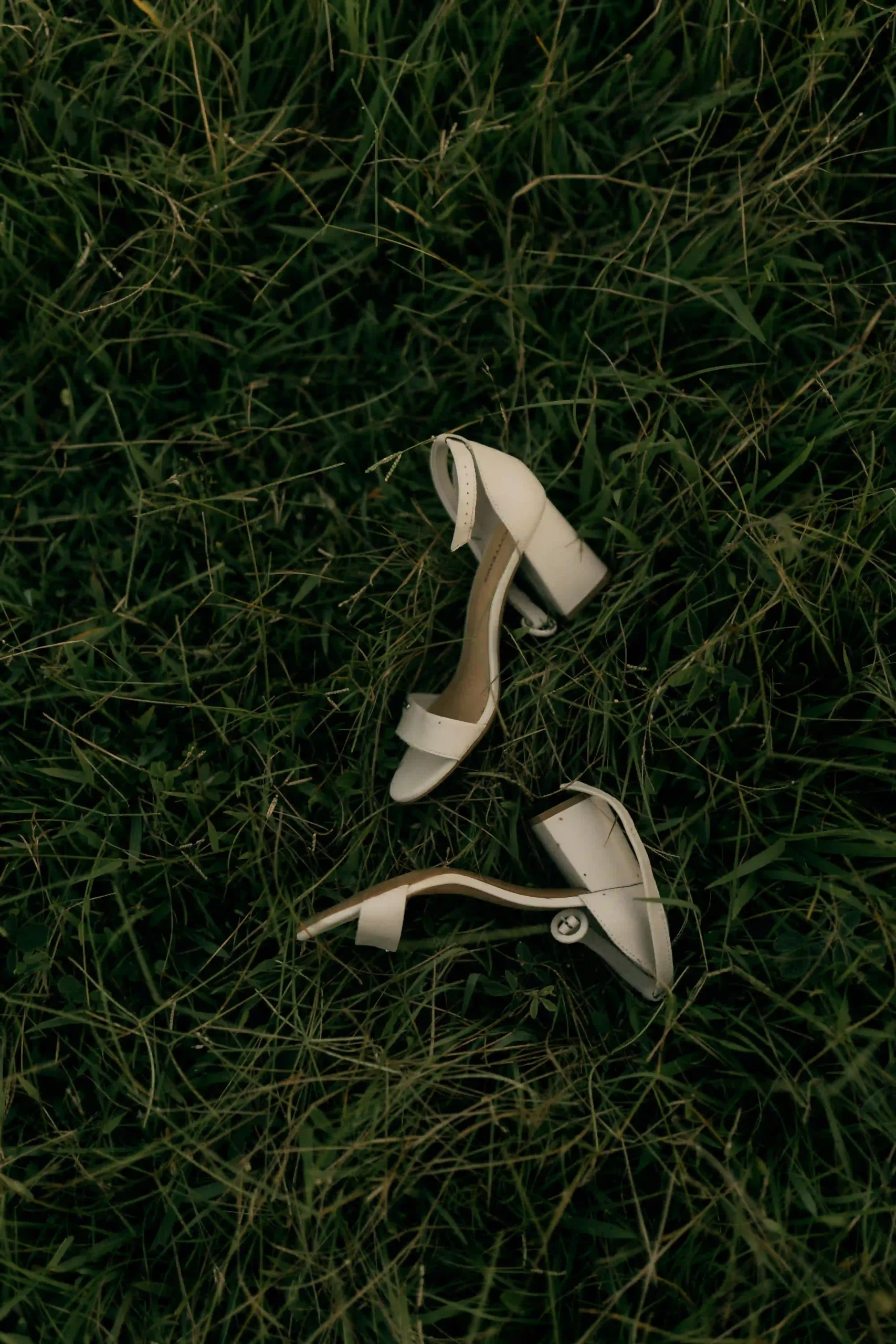 Pair of white block-heel sandals resting on green grass at an outdoor wedding