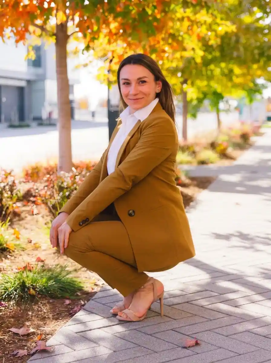 Woman in a brown coat and matching brown trousers sitting on a city walkway with autumn trees behind her