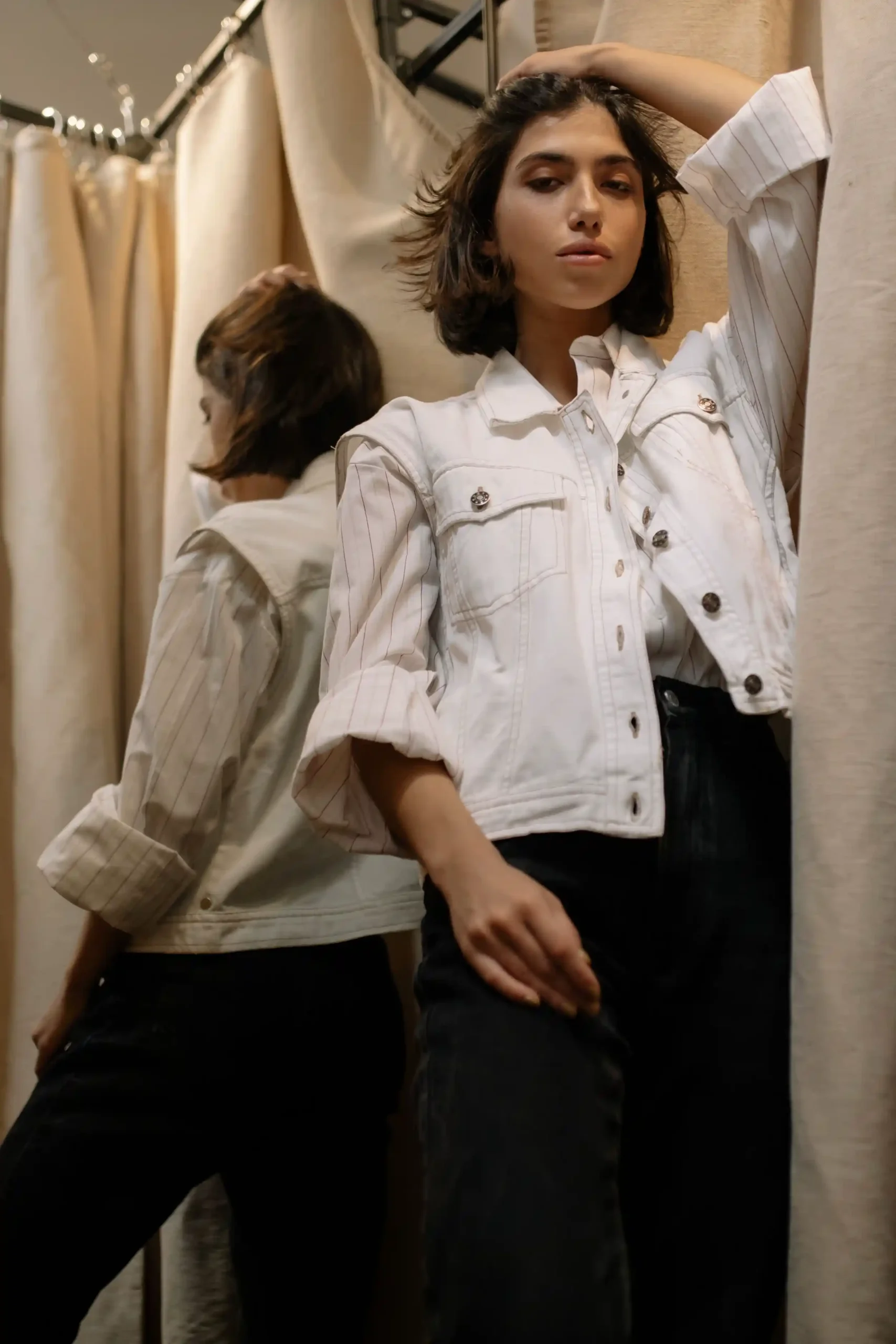A woman checking the fit of dark jeans and a white jacket in a fitting room mirror