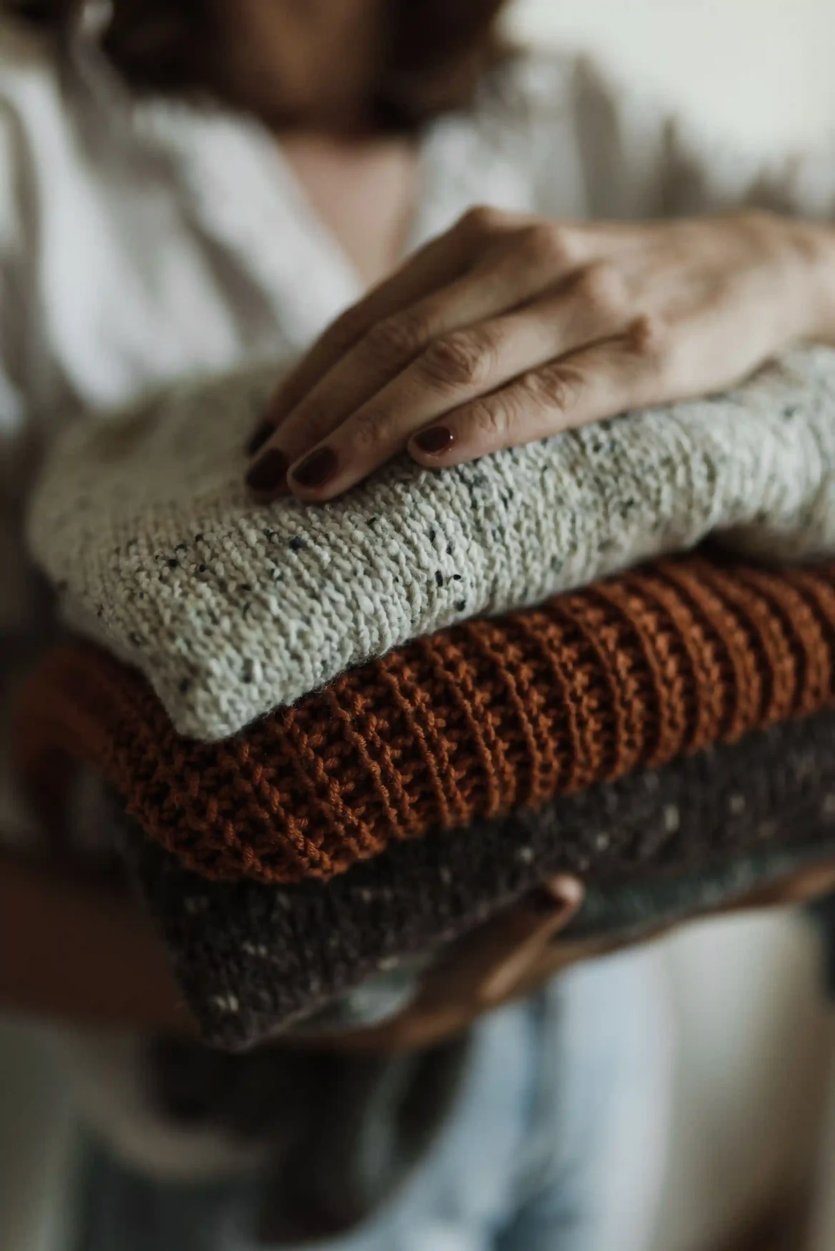 Hands holding a stack of folded knitted sweaters in neutral cream and beige tones