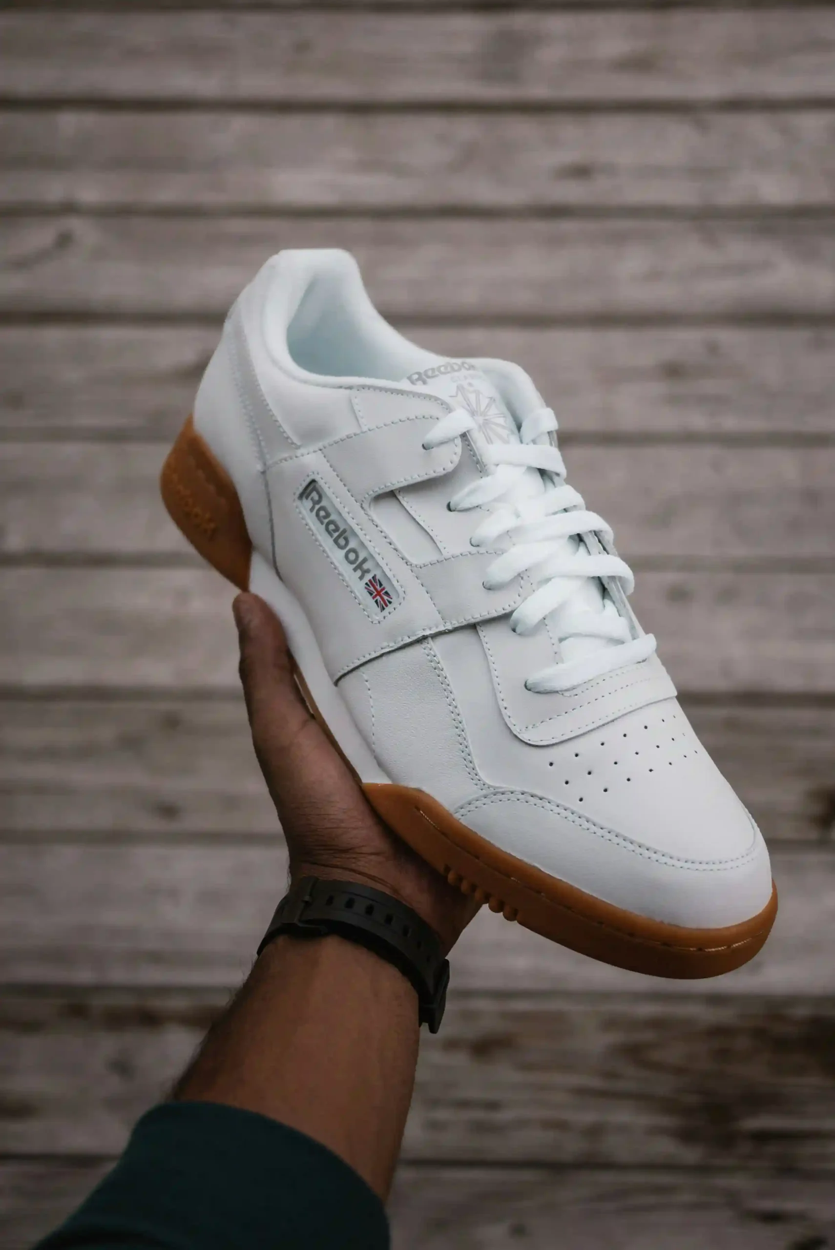 A low-profile white leather sneaker with a gum sole resting on a wooden floor.