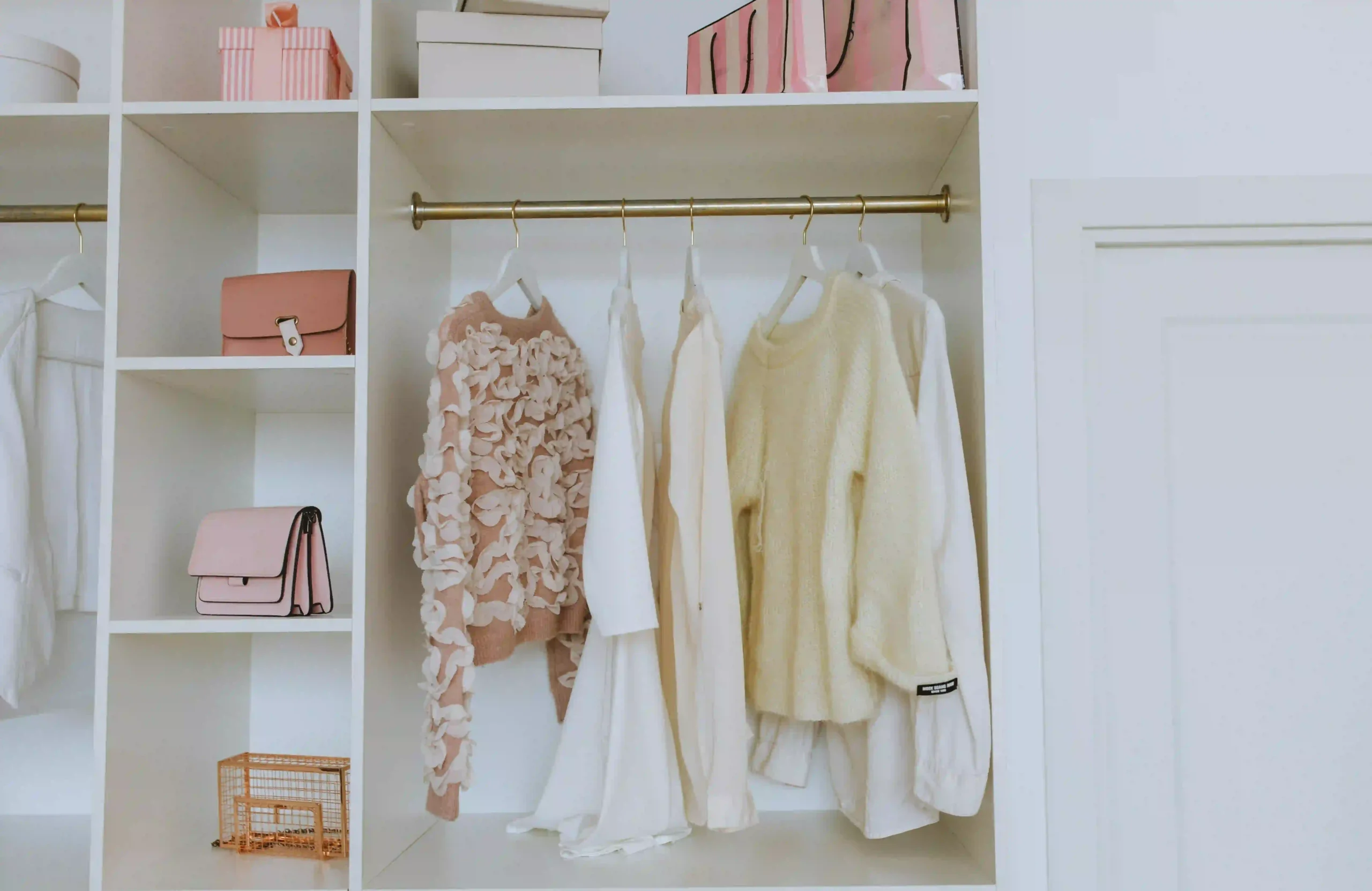 An organized white wooden closet holding a short rail of neutral-toned clothing with folded pieces on the shelf below