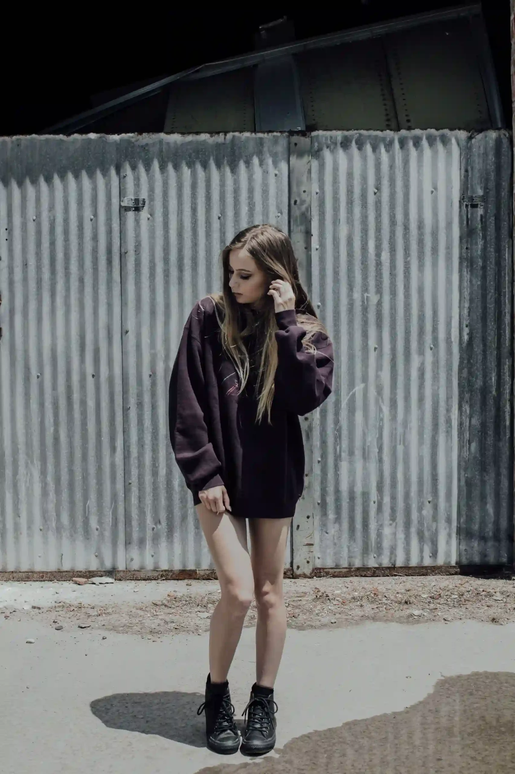 Woman in an oversized knit sweater and black boots posing against a corrugated metal fence, dark bookends holding a simple middle