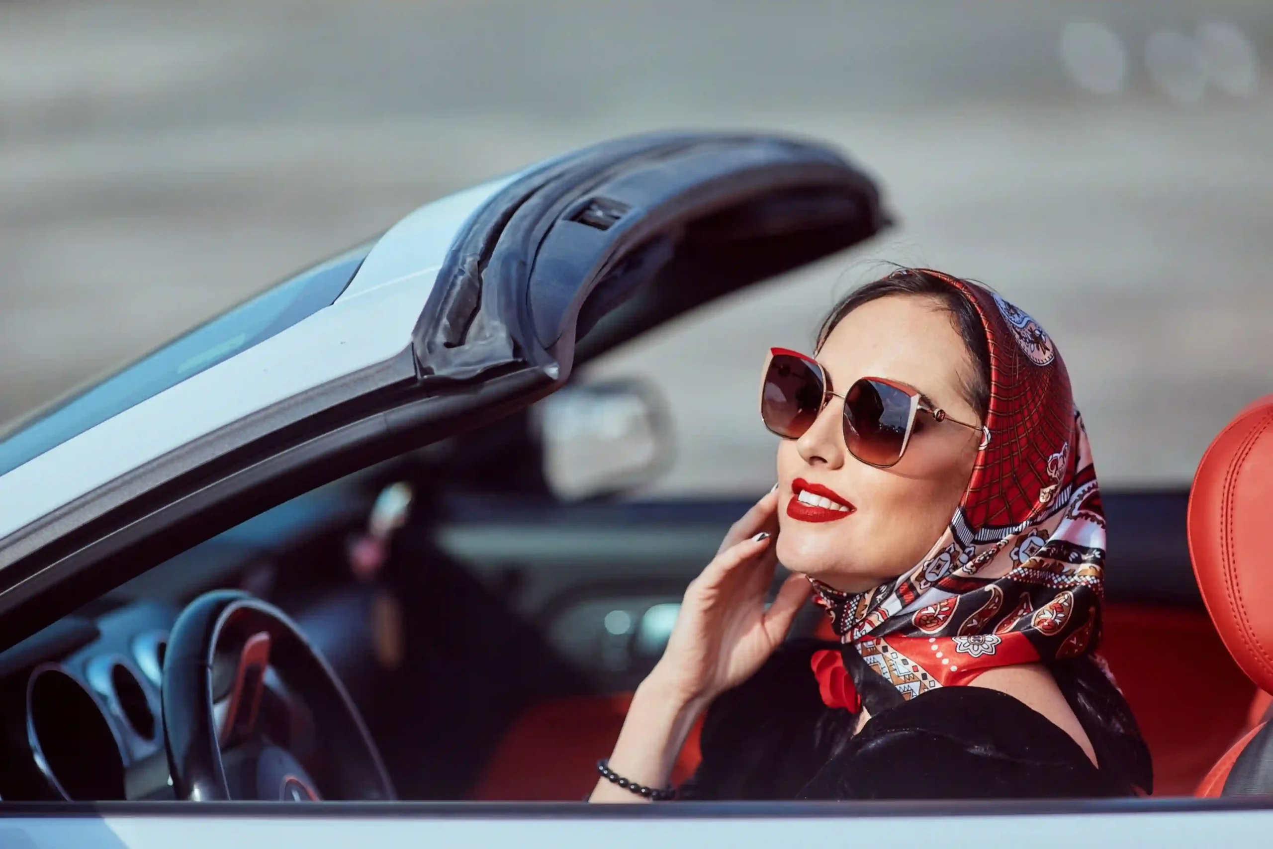 Woman wearing sunglasses and a silk headscarf tied under the chin, seated in a convertible on a sunny day