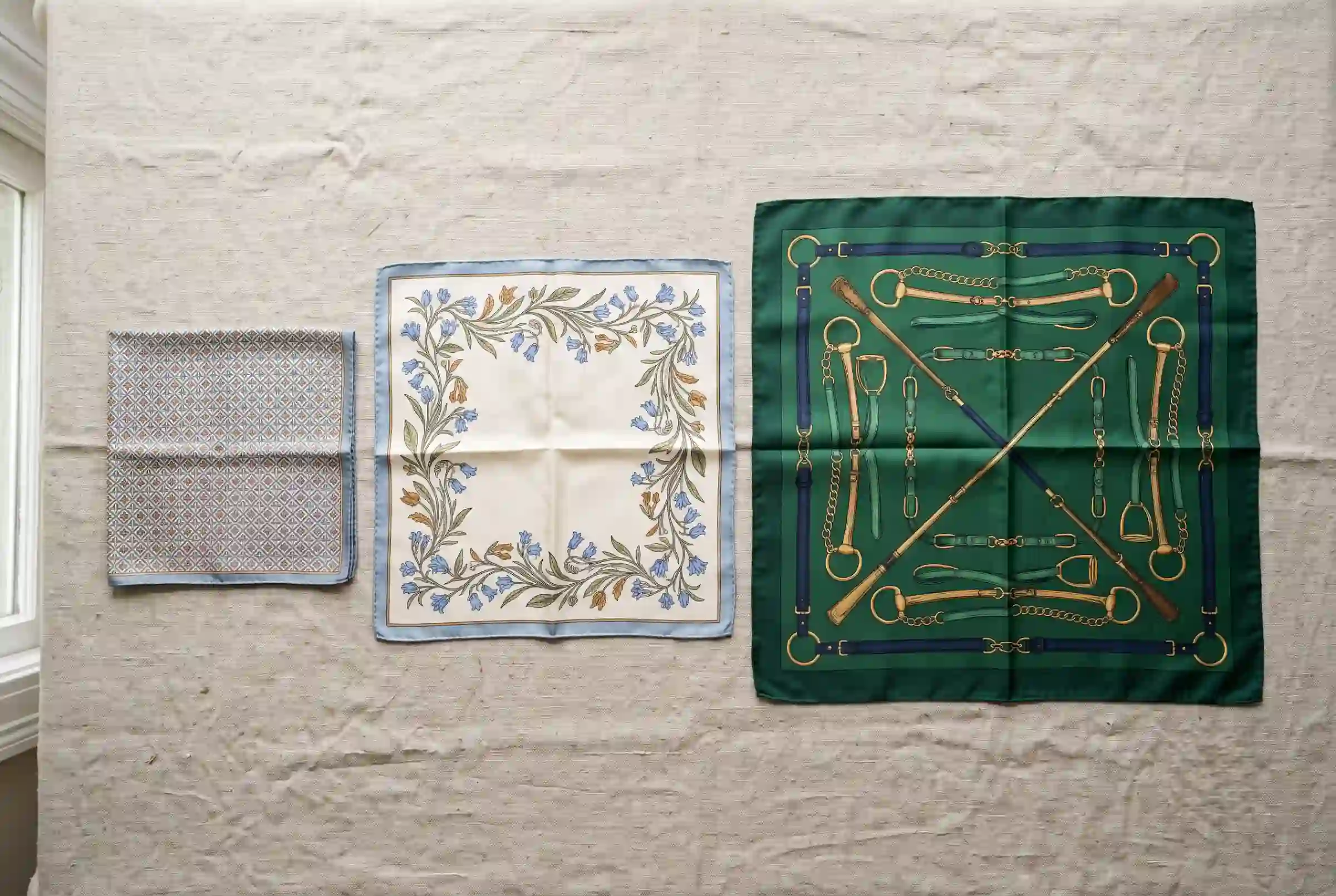 Three silk scarves in increasing sizes laid flat on linen fabric, small square, medium square, and large carré