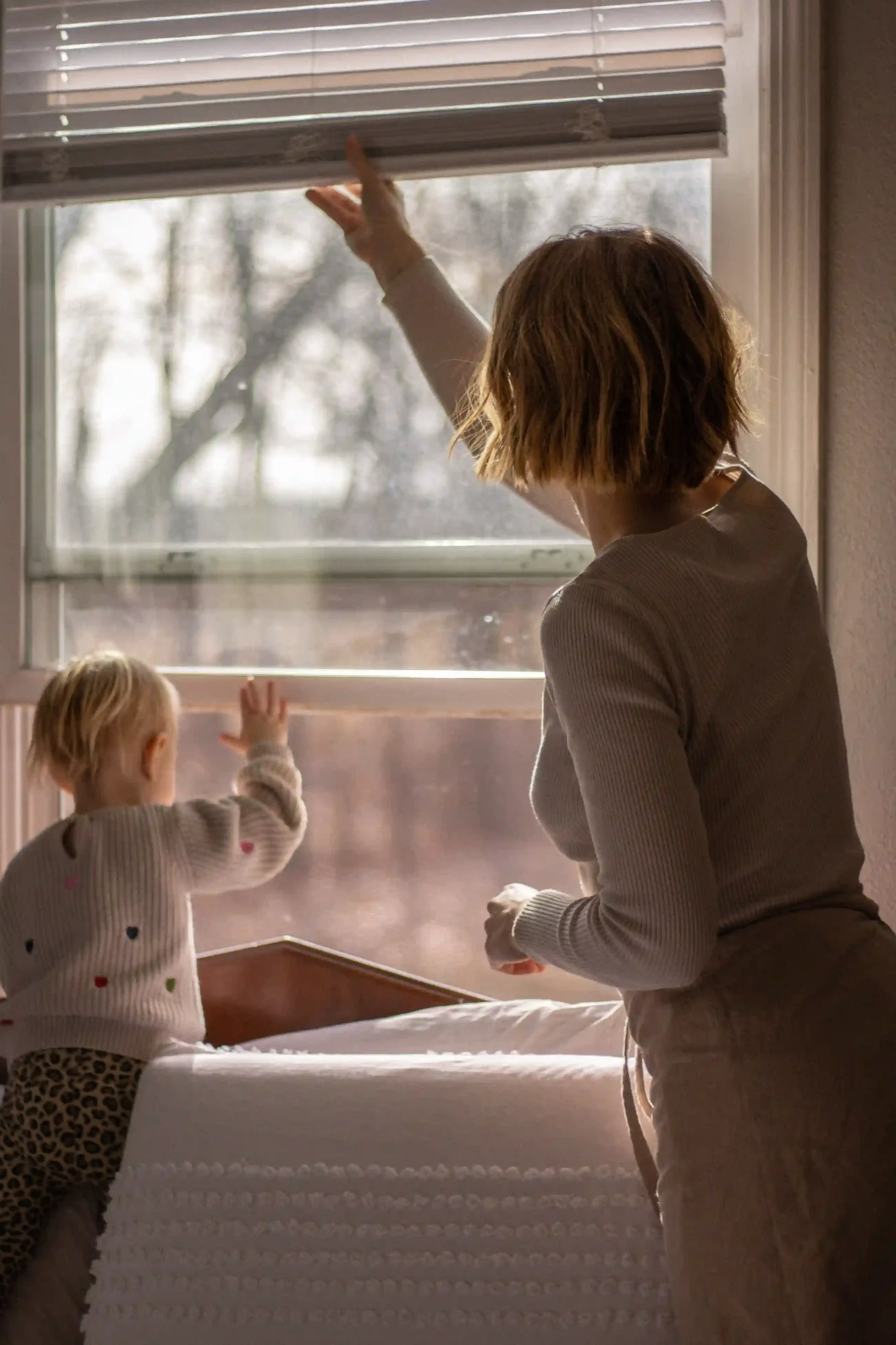A mother in a ribbed knit top and drawstring lounge pants adjusts the blinds while her toddler stands on the bed