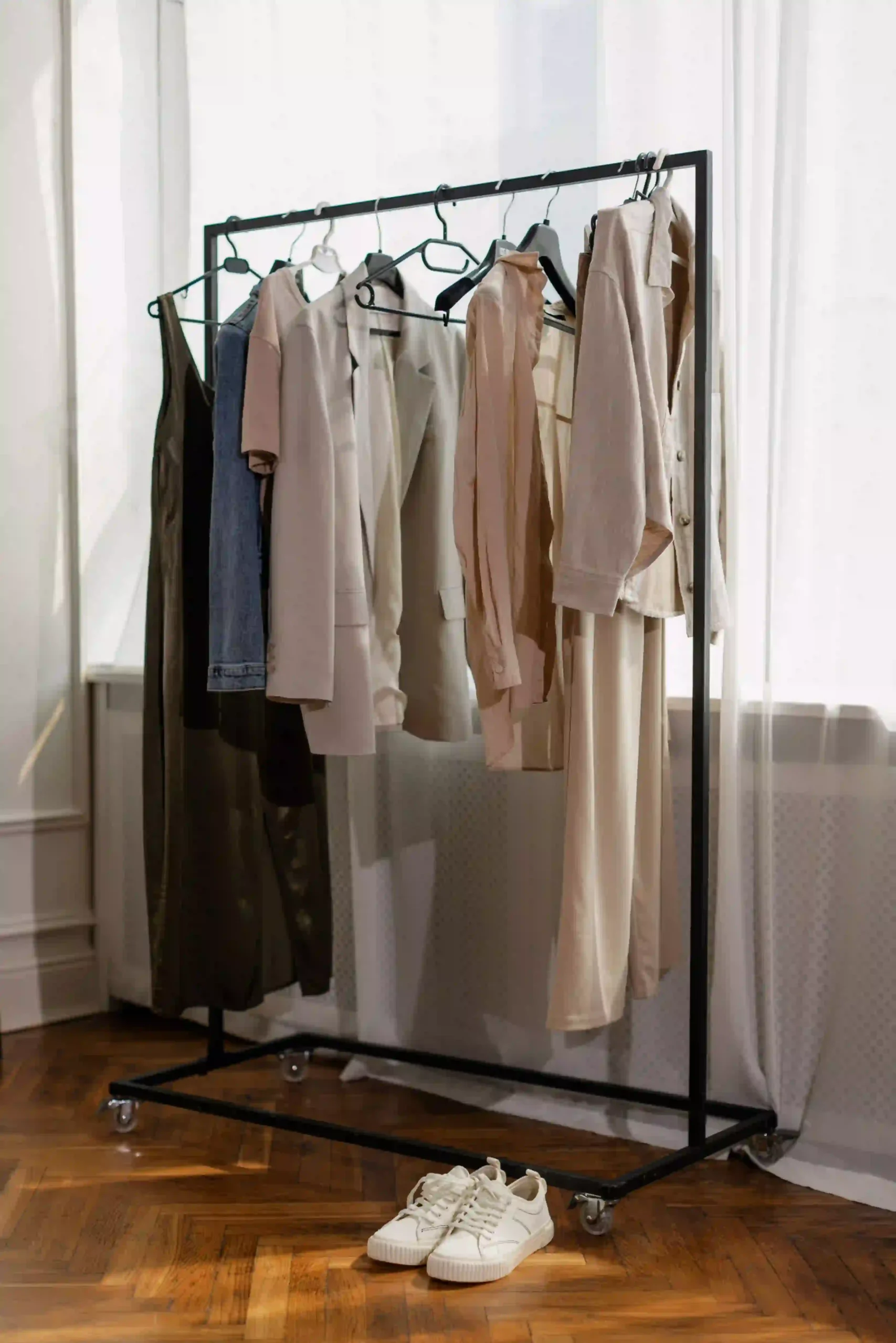 A minimalist clothing rack holding neutral-toned spring outfits with a pair of white sneakers on the shelf below in a bright room