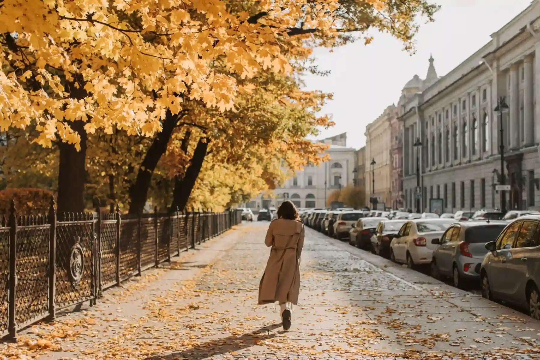 A woman in a beige trench coat walking on a leaf-covered city sidewalk in autumn